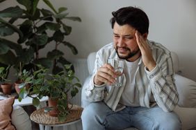 Young latino man wearing a striped button down and jeans is sitting on a couch at home with a headache. He has a pained expression and holds one hand to his temple while the other hand holds a glass of water. Houseplants sit beside and behind him.