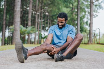 Man in a blue shirt, gray shorts, and black running shoes sitting on the ground in pain and holding thigh 