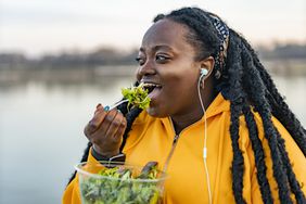 Young black woman with long locks is wearing a yellow sweatshirt and standing outside in front of a body of water. She is eating a salad and listening to music through corded earphones