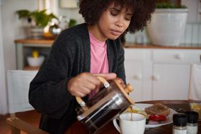 Black woman wearing a cardigan is sitting in her kitchen pouring coffee from a French press and having breakfast