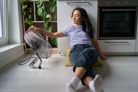 Tired overheated young Asian woman sitting on floor in kitchen near electric fan, cooling down at home during extreme summer heat.
