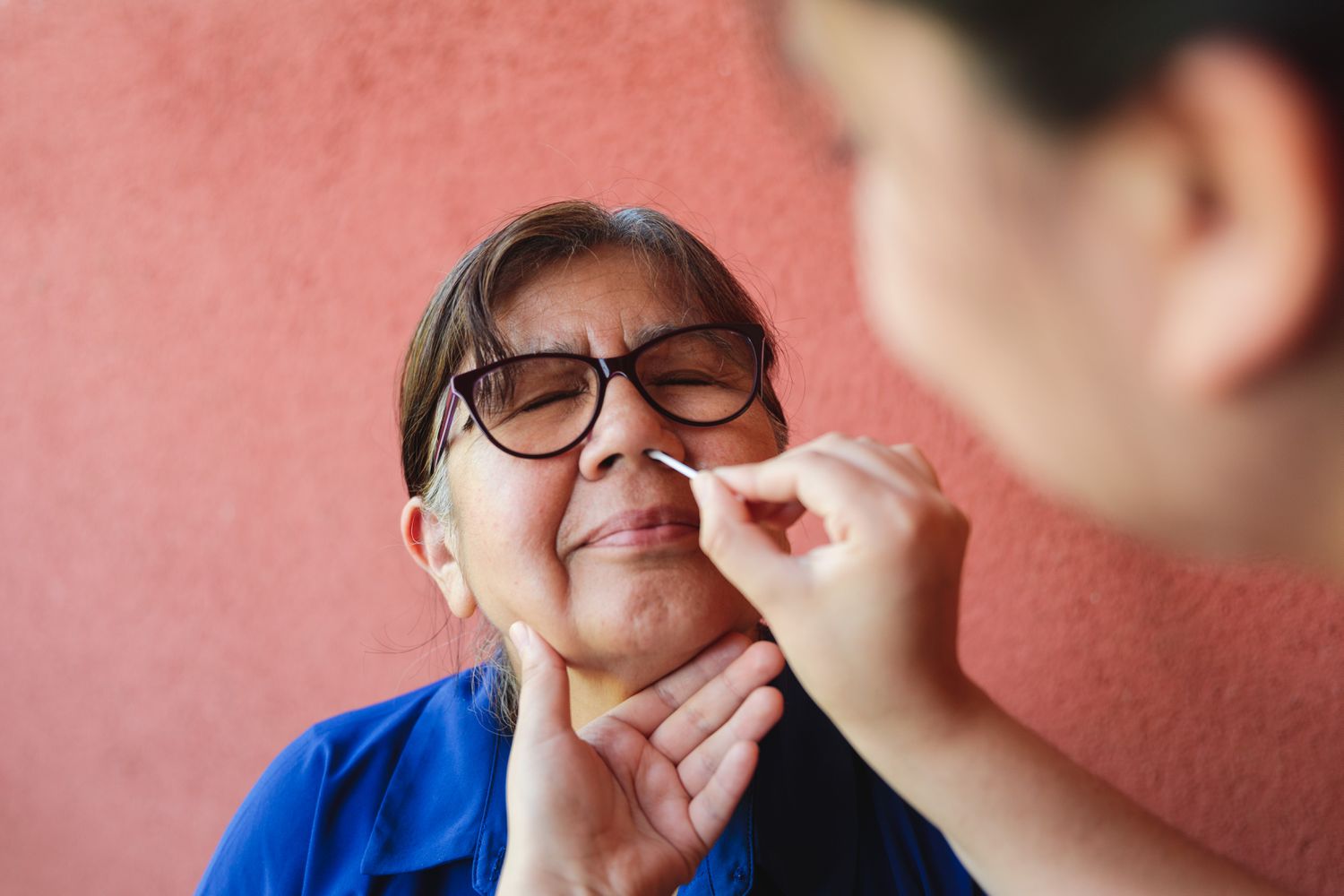 Older woman with glasses gets a nasal swab COVID-19 test. She's sitting in front of a salmon-colored wall. 