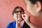 Older woman with glasses gets a nasal swab COVID-19 test. She's sitting in front of a salmon-colored wall. 