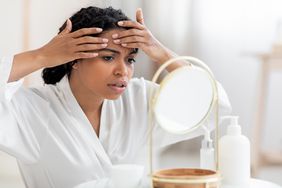 A woman puts her hands around a pimple on her forehead while looking in the mirror
