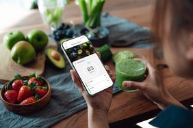 Woman looking at a calorie tracker on a smartphone, holding a green juice and sitting at a wooden table with strawberries, apples, avocados, and other fruits and veggies on the table