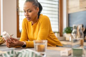 A Black woman wearing a yellow sweater is leaning over her kitchen counter and examining a supplement pill bottle. 
