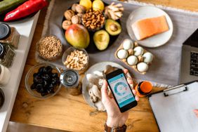 A person is holding a phone looking at a nutrition tracking app. On the table below is eggs, salmon, avocado, nuts, dried fruit, and more