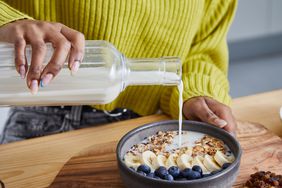 Close-up of a young woman's hands pouring a glass jug od milk over a bowl of cereal with bananas and blueberries. She wears a bright green sweater and has manicured nails