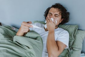 Young man in bed sick blowing his nose and checking his temperature. 