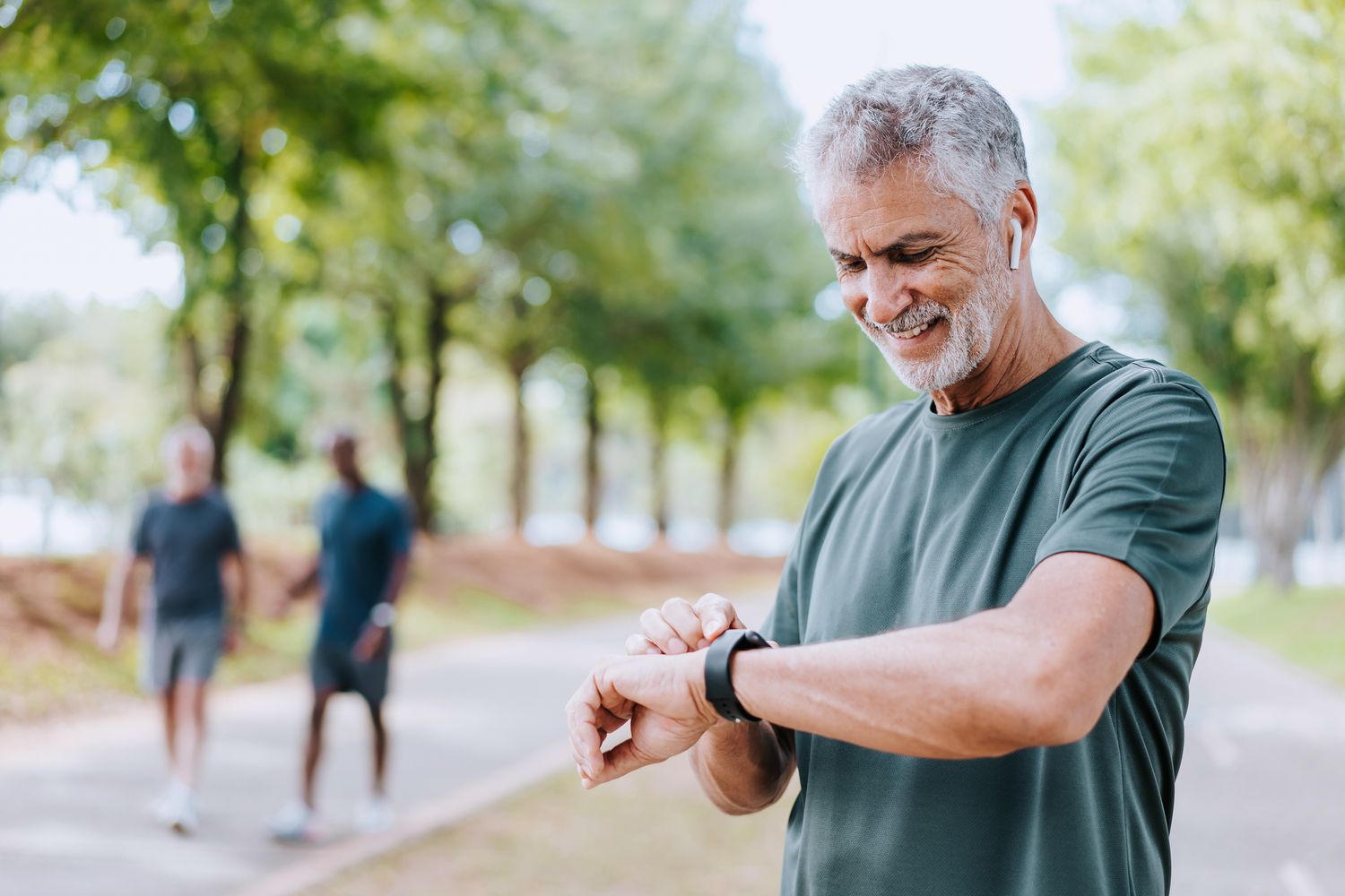 Older man wearing a sage green t-shirt is checking his smartwatch while outside on a walk. Two other men are walking in the background.