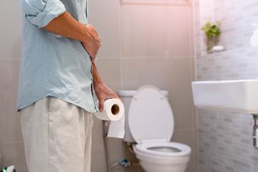 Close up of a man holding a roll of toilet paper and holding his stomach, in a bathroom setting with a toilet in the background