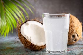 Glass of coconut water with split coconut in the back on a table