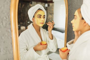 Woman wearing a white robe and white towel on her head is applying a yellow turmeric face mask to her face