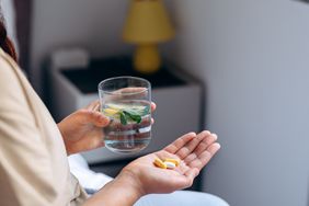 Close up of womans hands holding two types of supplements and a glass of water with fruit