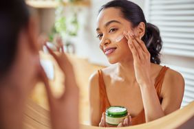 Young woman wearing an orange tank top is applying face cream in a mirror. She is holding the green jar of white skincare cream in her hand.