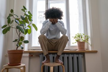 person with bipolar disorder sitting on window sill with feet up on stool holding head in sadness