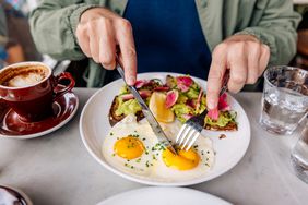 Close up of a man's hands using a fork and knife to cut into sunny-side up eggs, with avocado toast and coffee