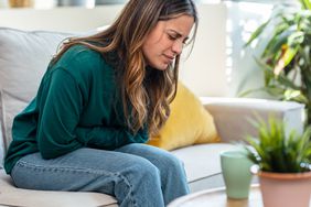 A woman sits on the couch and hunches over in pain