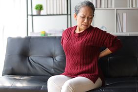 older woman sitting on a black couch wearing a red sweater and holding her lower back in pain