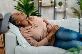 A woman lies on her couch while holding her stomach in pain