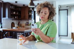 Middle-aged black woman with light curly hair wearing a gree tshirt and sitting in her kitchen; she is looking at a smartphone and holding a prescription pill bottle