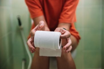 A person sits on a toilet while holding a roll of toilet paper