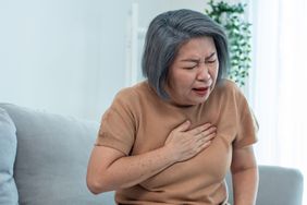 older woman experiencing heart attack sitting on couch grabbing her chest in pain