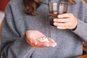 Postbiotic Supplement: A close up of a woman wearing a gray sweat holding a glass of water and several supplement capsules