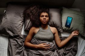 Young black woman wearing a gray tank top is lying in bed (gray sheets and bedding). She is on her back, awake and looking up. A smartphone sits on a pillow beside her, showing an alarm icon. 