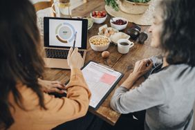 dietitian explaining nutrition facts on laptop to person while sitting on a wooden table with bowls of different foods