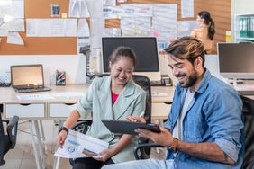 two people sitting in office chairs laughing while looking at an ipad