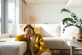 woman in yellow sweater holding head and leaning on couch while sitting on the floor