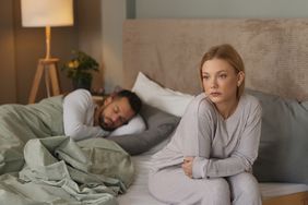 Woman sitting on bed, looking unhappy, with male partner behind her in bed asleep
