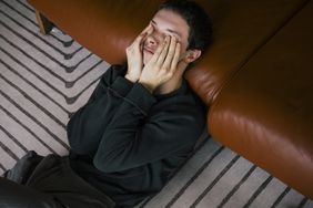 frustrated person lying on the floor with head on couch covering their eyes with their hands