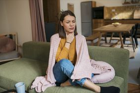 White woman with short brown hair holding her sore throat. She sits on a green couch and wears jeans, a mustard shirt, and a pink blanket around her shoulders