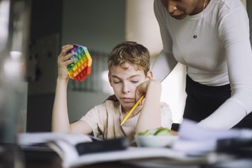 A boy uses a fidget toy and holds a pencil while trying to do schoolwork. A woman is pointing to the paper.