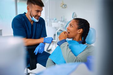 Young black woman sits in a dentist office chair. She points to her tooth while talking to a white male dental technician.