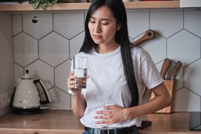 Sick young Asian woman standing in kitchen holding glass of water suffering from stomach ache, unhealthy girl feeling unwell during period, touching belly stomach having menstrual cramps