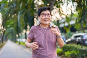 Older Asian woman with short hair, wearing a pink tshirt and red-rimmed glasses, is walking outdoors, listening to music through wired headphones. She wears a smartwatch to track step count
