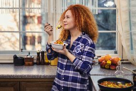 A woman stands in her kitchen eating a bowl of food she cooked