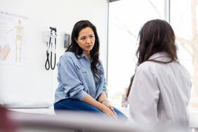 Asian woman wears a striped button down and jeans, sitting in a doctor's office. She is listening attentively to the female doctor