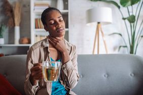 A young black woman holds her throat in pain while holding a cup of tea. She sits on a gray sofa in a living room at home.