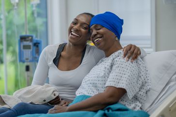 A woman with cancer wears a head covering and hospital gown as she sits with her daughter