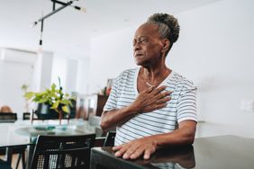 older woman standing in the kitchen holding on to the table in pain while clutching her heart
