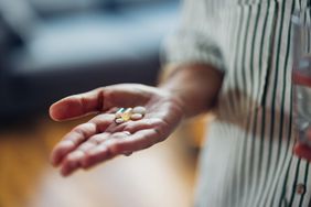 A close-up shot of a person's hand holding various pills and medication. They are wearing a green striped button down shirt
