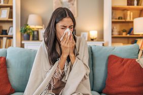 Woman with brown hair and tan skin sitting in a living room, on a blue couch, wrapped in a tan blanket, blowing her nose into a tissue