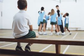 A boy sits on the side alone as his peers play a game
