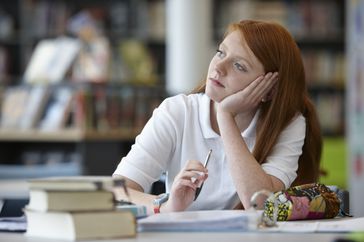 teenager with red hair and white shirt sitting on a desk feeling distracted and looking away from homework