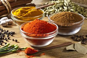 Three glass bowls filled with ground spices—colors of orange, yellow, and brown—are arranged on a rustic wood table. Other spices like black pepper, nutmeg, cinnamon sticks, cardamom, clove and bay leaf are around the bowls