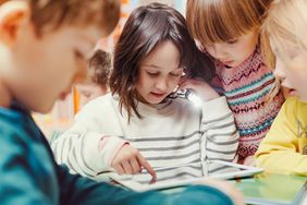 A little girl touches her ear with a group of other little kids surrounding her as they look at a tablet